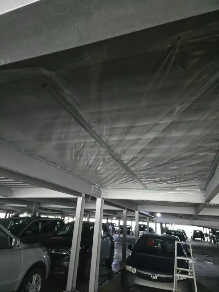 Parking couvert avec plusieurs voitures garées ; le plafond est enveloppé d'une feuille de plastique transparent et un escabeau blanc est placé près d'un véhicule.