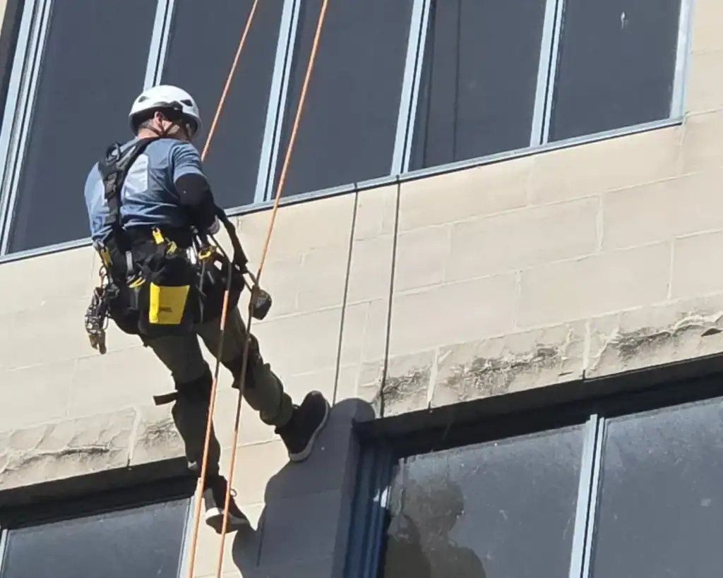 Un travailleur portant un équipement de sécurité est suspendu à des cordes pour nettoyer les fenêtres extérieures d'un bâtiment.