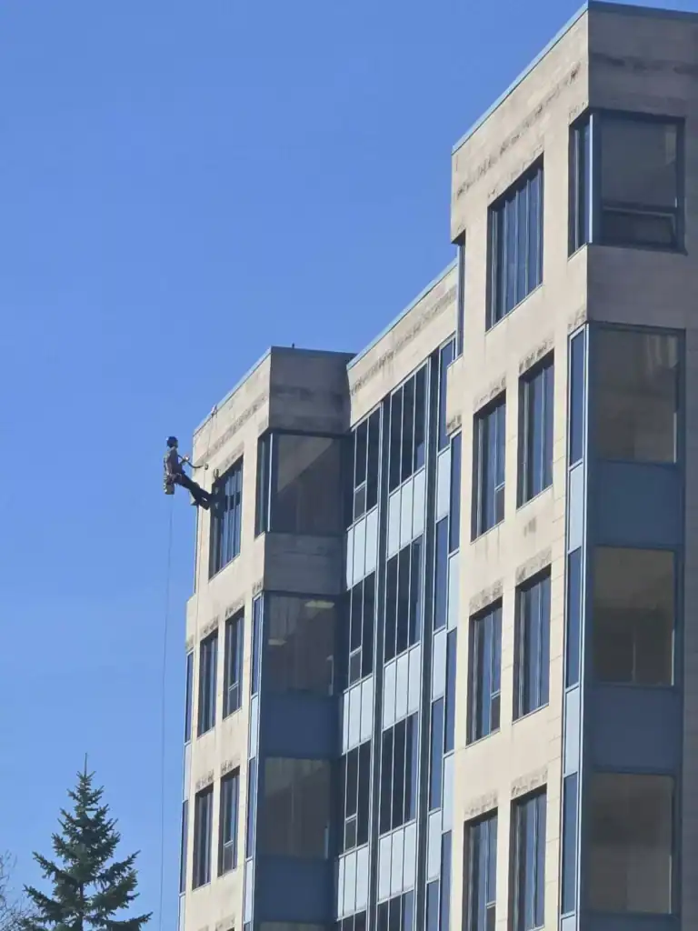 Un travailleur est suspendu à une corde, nettoyant les fenêtres à l'étage supérieur d'un bâtiment à plusieurs étages sous un ciel bleu clair.