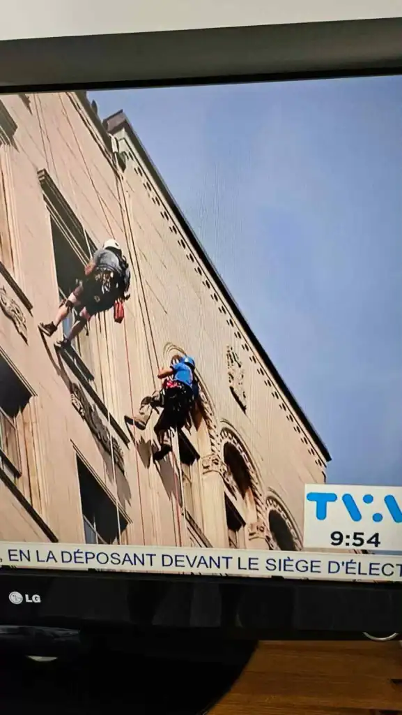 Deux ouvriers portant des équipements de sécurité sont suspendus à des cordes, effectuant des travaux d'entretien ou de nettoyage sur le côté d'un grand bâtiment en pierre. Une bannière de journal télévisé est partiellement visible en bas.