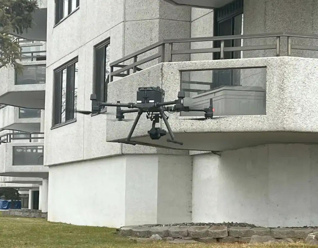 Un drone noir équipé d'une caméra plane près du balcon d'un immeuble d'habitation blanc et moderne, au-dessus d'une zone herbeuse.